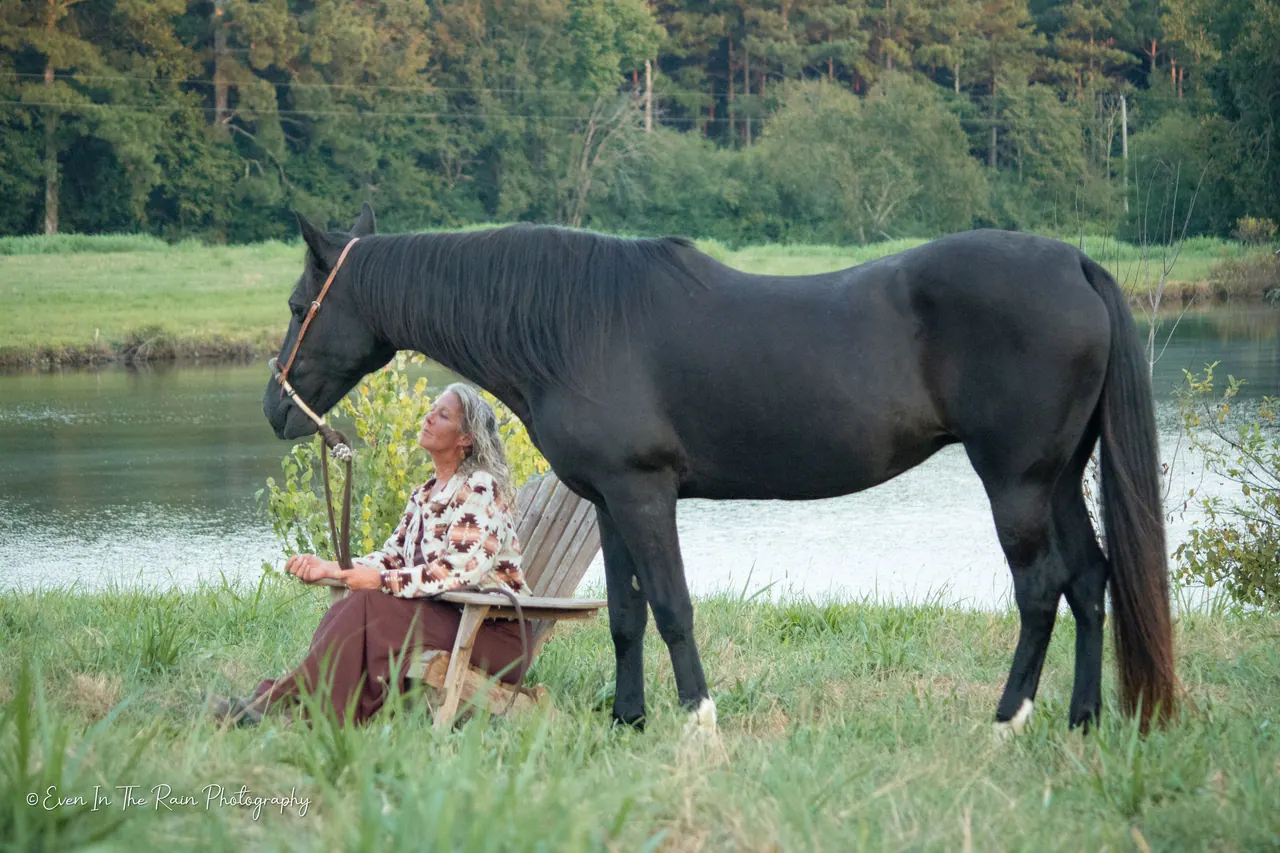 Leslie sitting quietly in an Adirondack chair beside her black horse by the lake